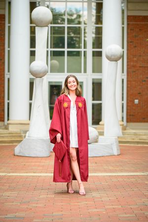 Graduate in red Elon graduation gown poses in front of campus building with decorative white pillars.