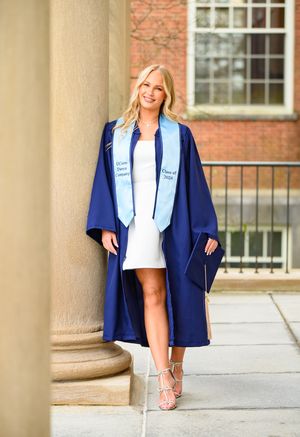 Academic graduation portrait in navy blue gown and stole against brick building.