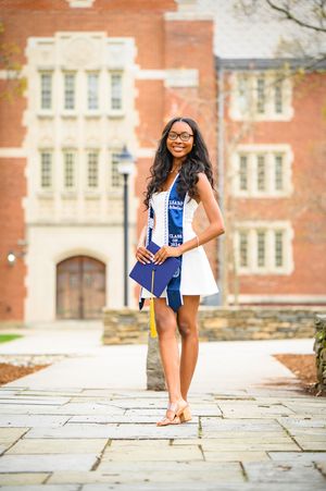 Graduate in white dress and blue stole poses on stone pathway in front of historic brick campus building.