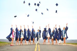 Graduates in blue robes toss caps in the air while celebrating on an outdoor road.