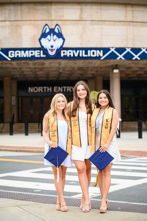 Three graduates in gold stoles pose with grad caps outside Gampel Pavilion entrance.
