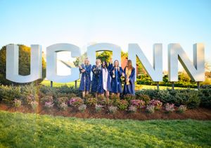 Graduates in blue robes pose together in front of large UCONN letters on campus lawn.