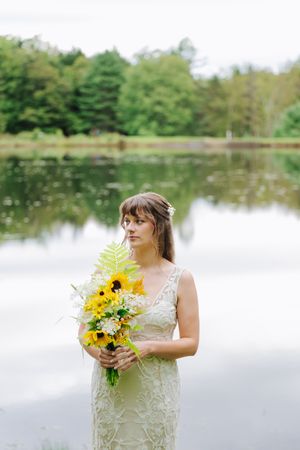 A bride in a lace dress holds a vibrant yellow sunflower bouquet while standing by a serene lake with green trees.