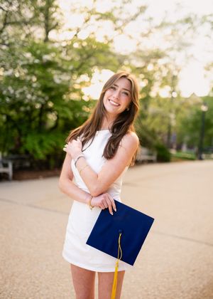 A graduate in a white dress holds a blue graduation cap with gold tassel while standing outdoors during golden hour.