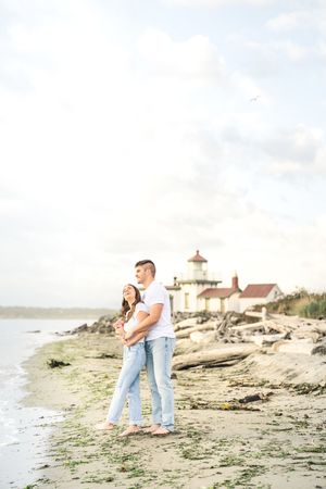 Couple shares tender moments during sunset photoshoot on rocky beach with lighthouse in background.