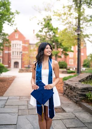 Graduate in a white dress and blue graduation stole stands smiling on a stone pathway with red brick buildings in background.
