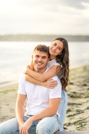 A couple in white t-shirts and jeans embrace on a beach at sunset with water and mountains in the background.