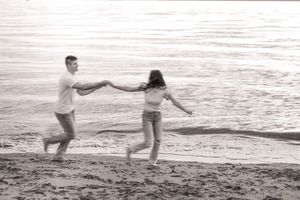A couple walks along a beach at sunset while holding hands in a romantic black and white scene.
