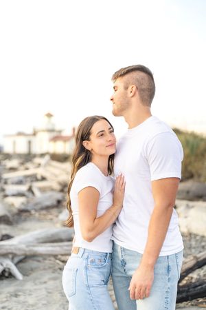 A romantic couple in matching white t-shirts and jeans embraces on a sandy beach at sunset.