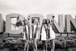 A black and white photo of graduates celebrating in front of large block letters on a college campus.