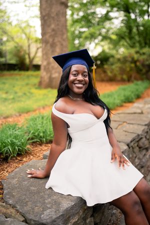 Graduate in blue cap and white dress sits on stone wall during outdoor graduation photo session.
