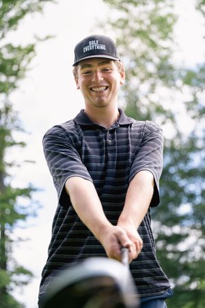 A golfer wearing a striped polo shirt and baseball cap smiles while gripping a club on a sunny day.