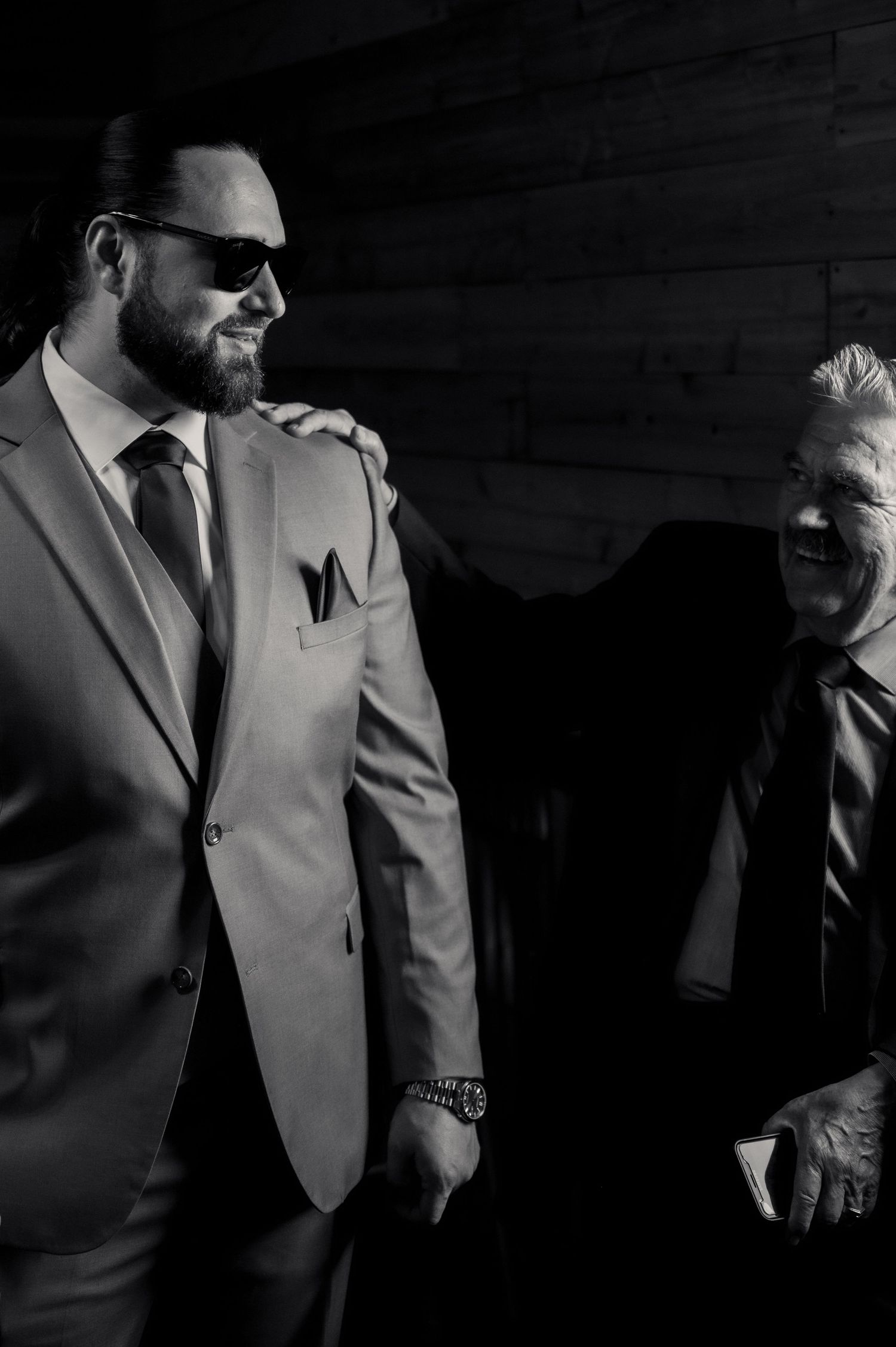 Two men in suits share a candid moment in dramatic black and white lighting.