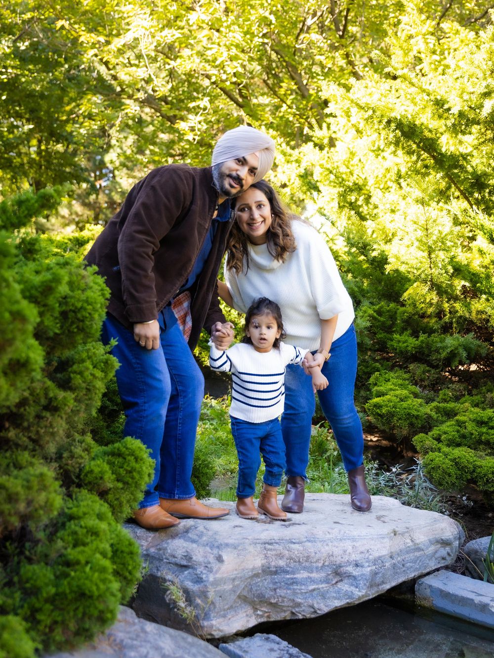 Family of three posing on a rock in a lush green garden, parents holding a toddler girl wearing a striped shirt.