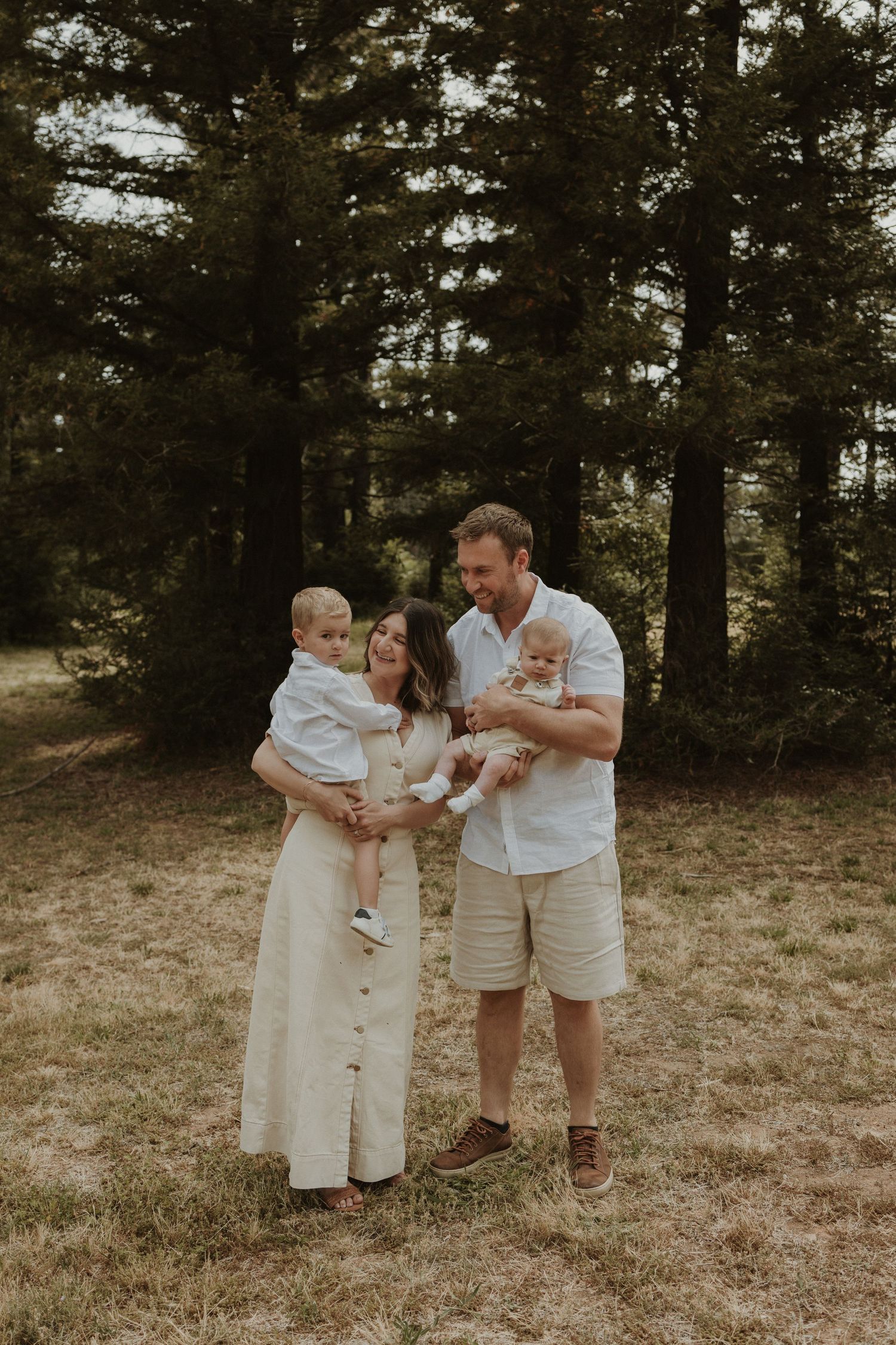 Family stands together in Pialligo Redwood Forest at sunset, wearing light colored clothing in a natural outdoor setting.