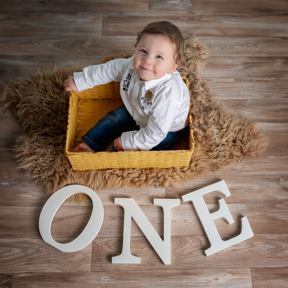 First birthday milestone photo with baby sitting in yellow wooden box surrounded by fluffy brown rug and white ONE letters.