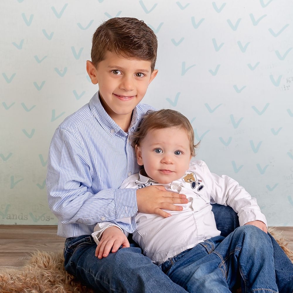 Two children in coordinating blue and white outfits pose together on a light colored floor against a patterned wall.