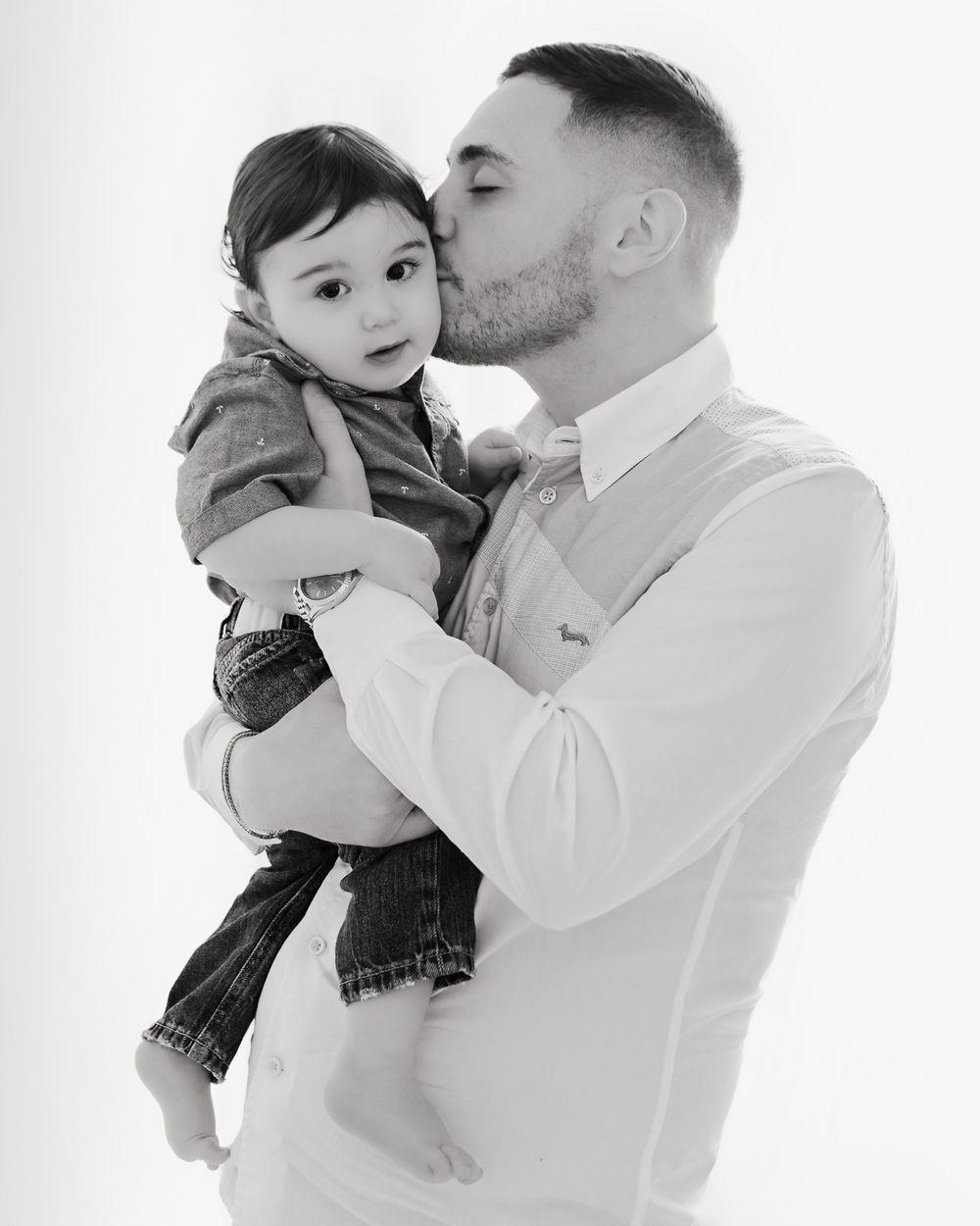 A tender black and white portrait showing an affectionate moment between an adult and baby in formal attire.