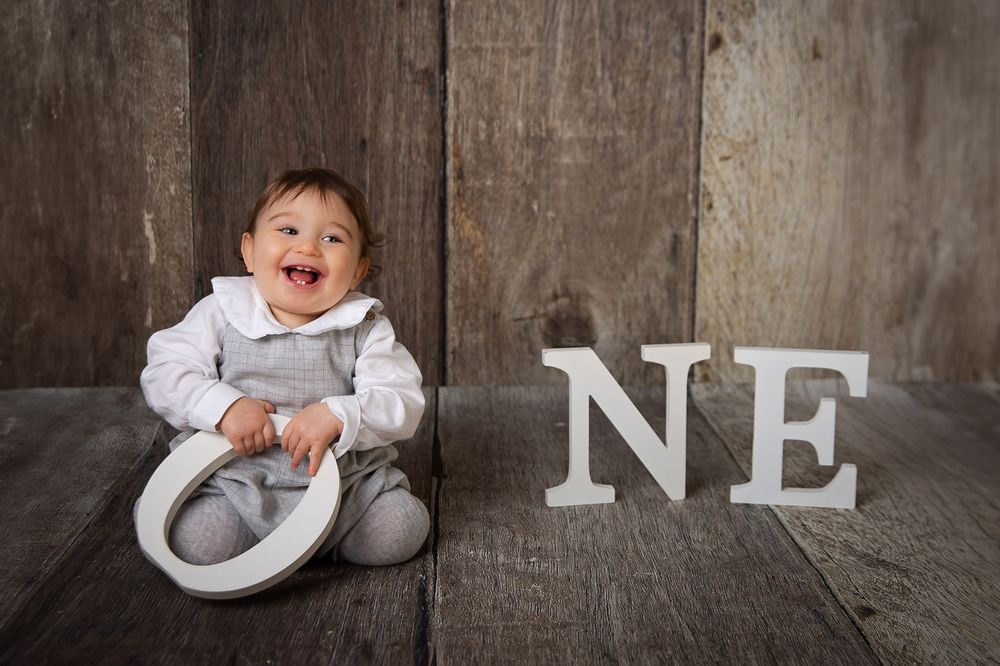 Baby in white sweater sits and plays on concrete floor while making playful expressions.