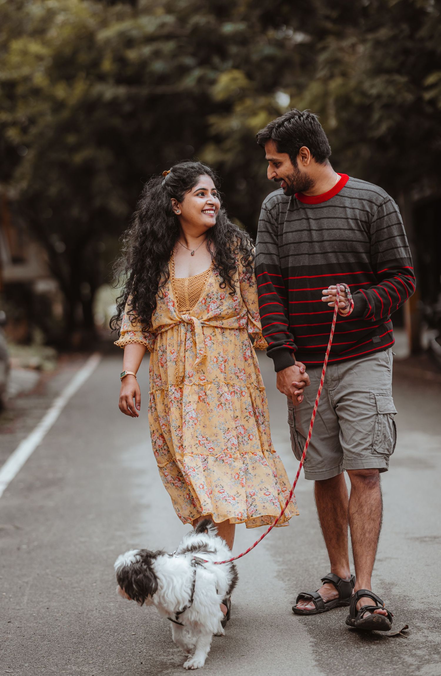 Couple walking their small white dog on a tree-lined path.