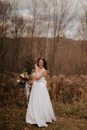 Mariée souriante dans un champ aux teintes automnales, robe blanche fluide et bouquet rustique.