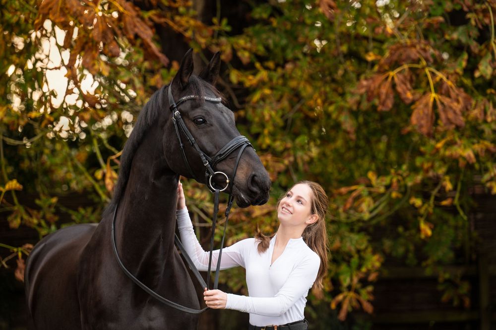 Horse standing beside her handler in an autumn landscape with golden leaves, captured in soft natural light.