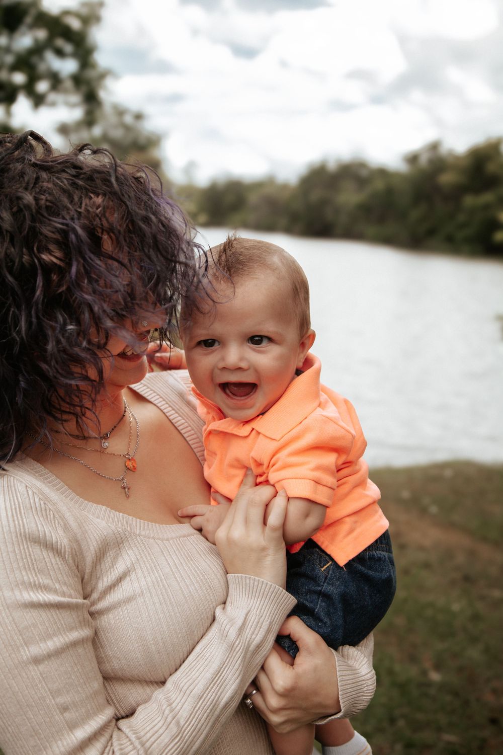 A baby wearing bright orange laughs while being held near a waterfront setting.