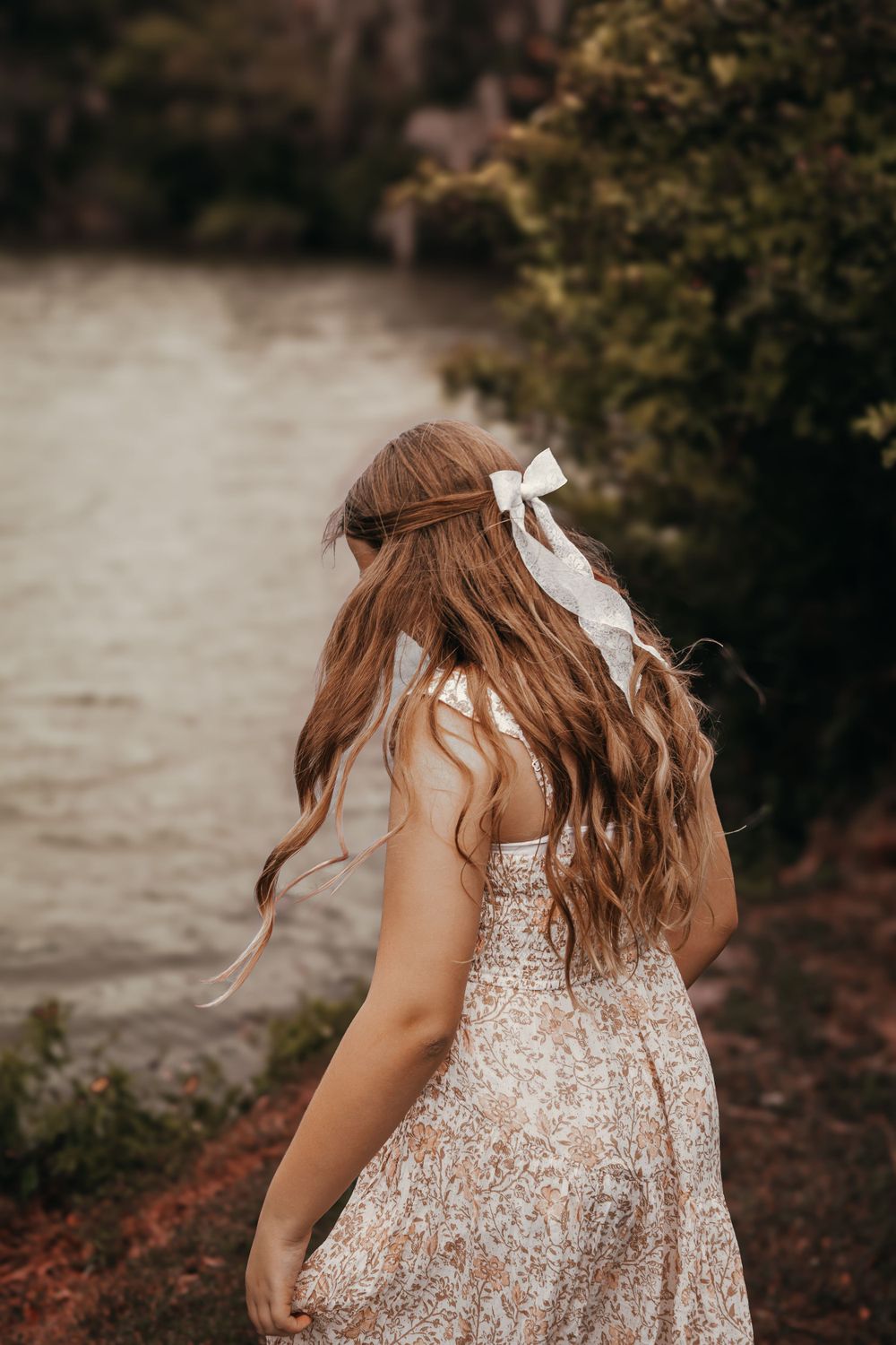 Back view of person in floral dress with long wavy hair and white ribbon bow by lake.