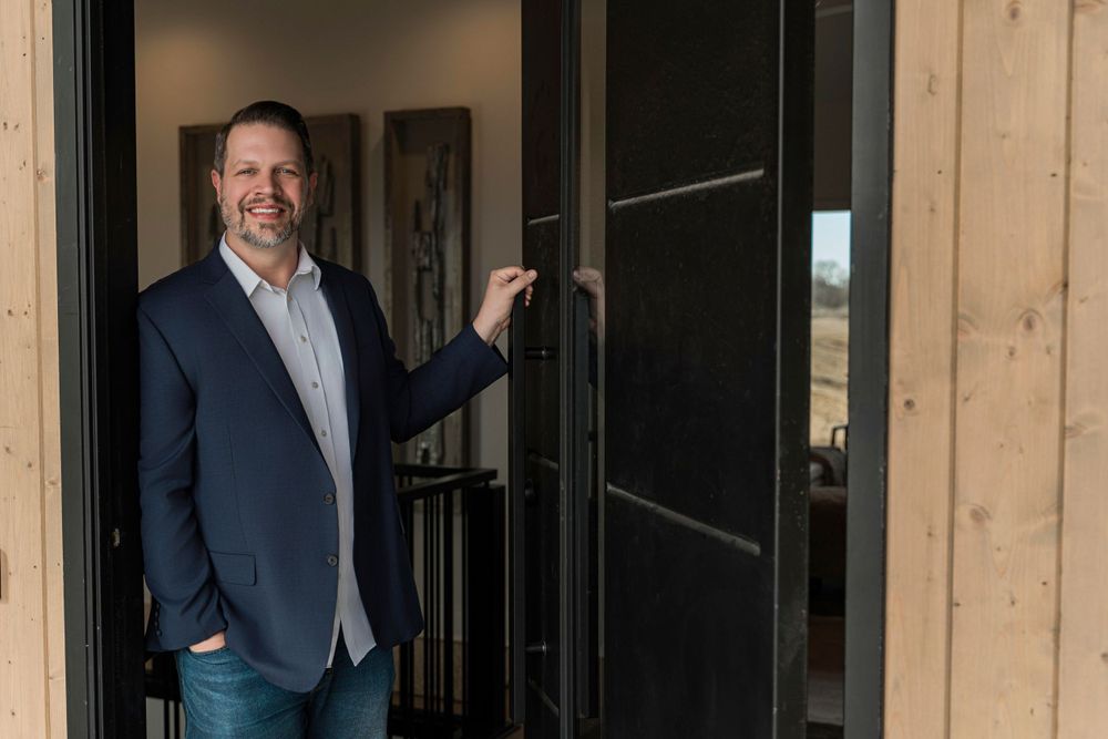 Person in navy blazer and jeans stands smiling by modern black door on wood-framed building exterior.