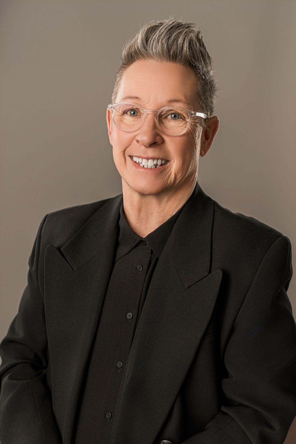 Professional headshot of an individual in a black suit and dark button-up shirt wearing clear glasses against a neutral background.