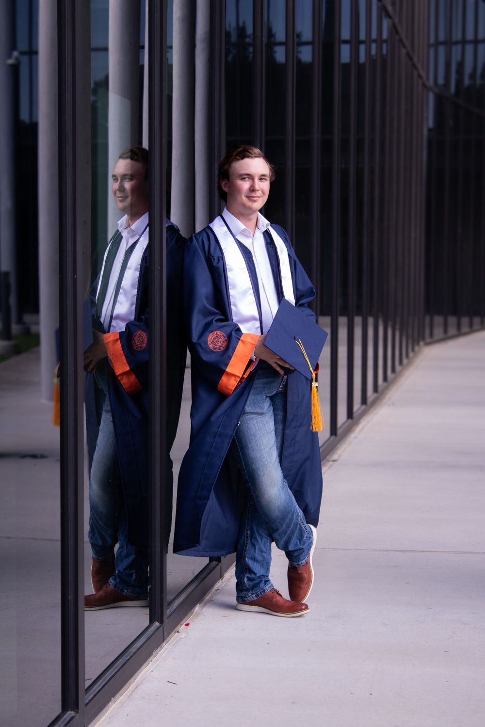 Graduate in blue cap and gown holding orange folder stands against reflective glass building exterior.