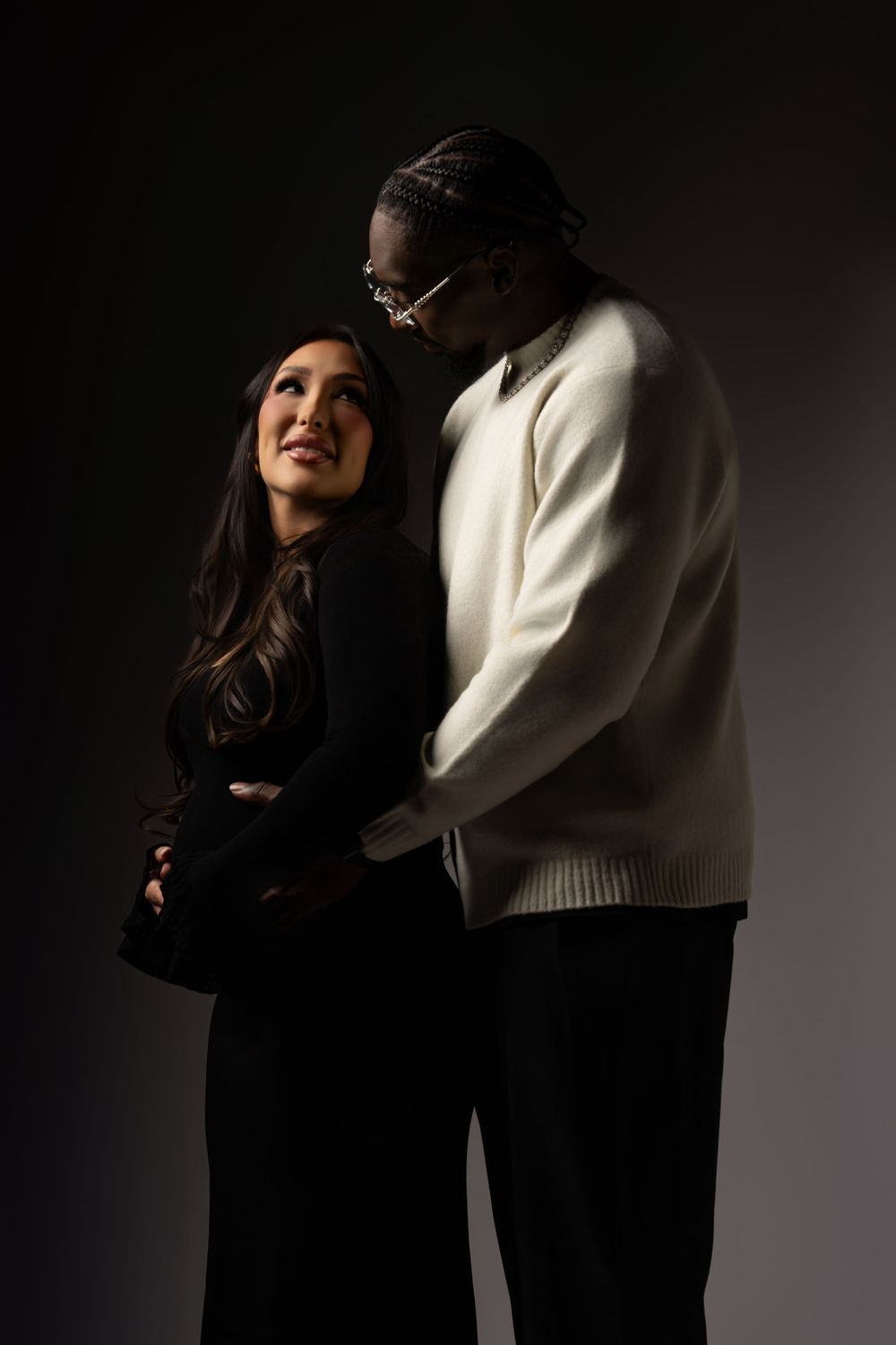 A couple embraces intimately in a dark studio setting with dramatic lighting and contrasting black and white outfits.