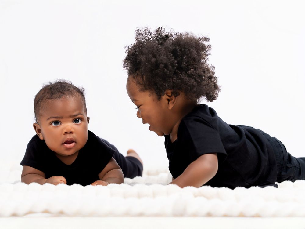 Two babies in black outfits lying on a white carpet while looking at each other.