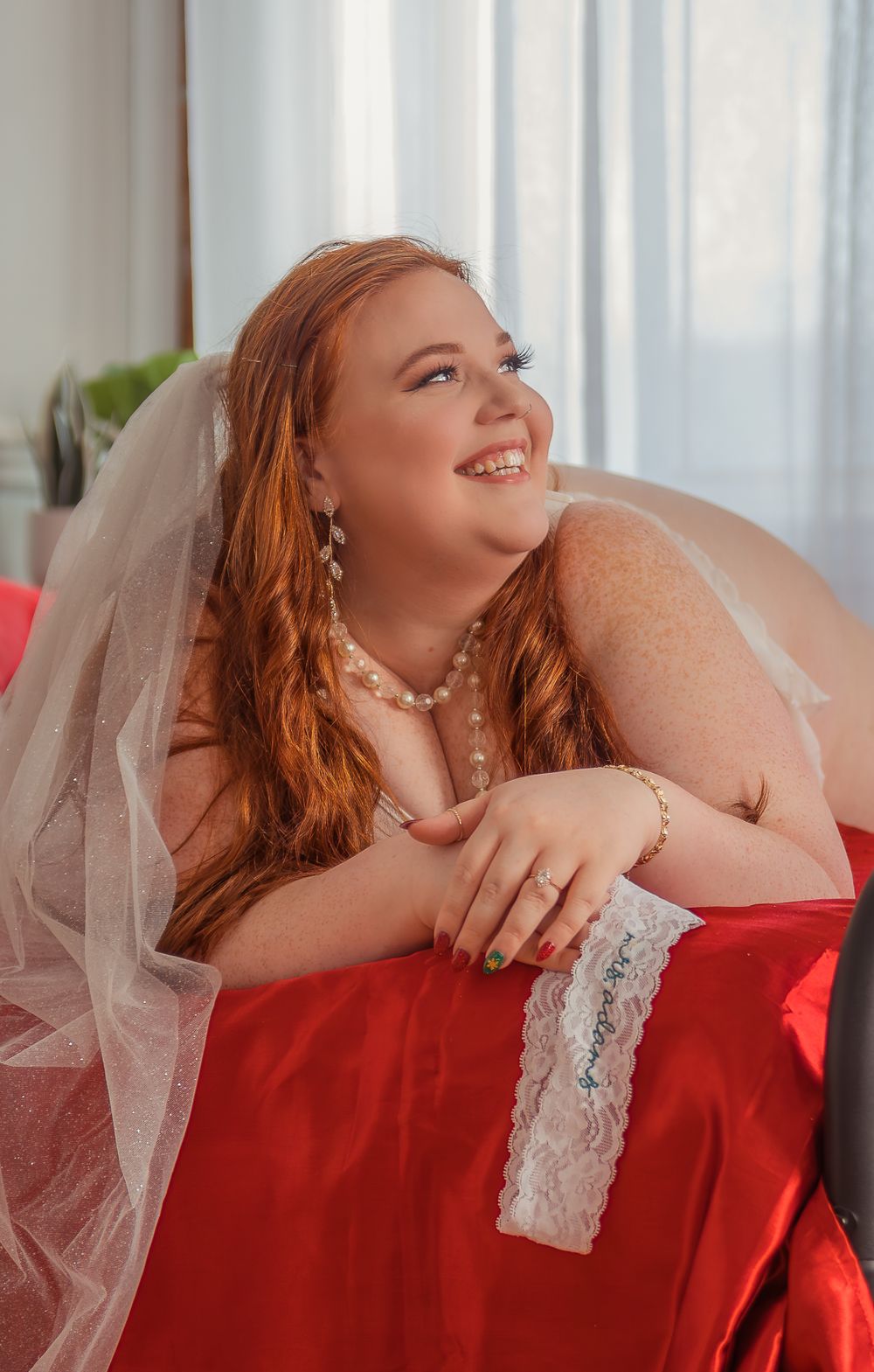 A bride wearing a red dress and veil smiles while reclining on red bedding with lace trim.