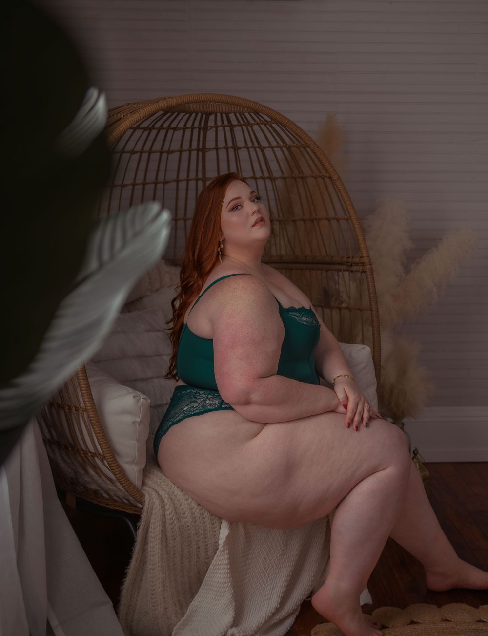 Moody boudoir photo of model in emerald lingerie seated on rattan egg chair against white backdrop.