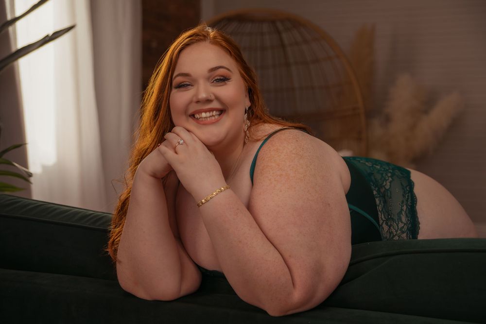 A person with red hair smiles warmly while relaxing on a dark green couch in a sunlit room.