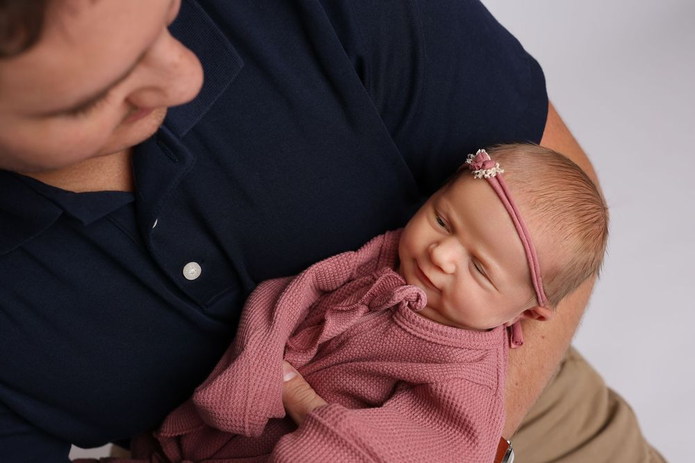 A father in a navy shirt tenderly holds his sleeping newborn baby wrapped in a pink swaddle with a floral headband in Covington, GA