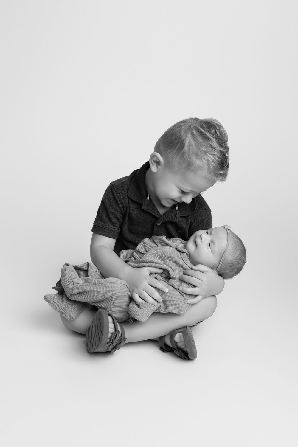 Black and white photo of a young boy sitting cross-legged, lovingly cradling a newborn baby in his arms in Covington, GA