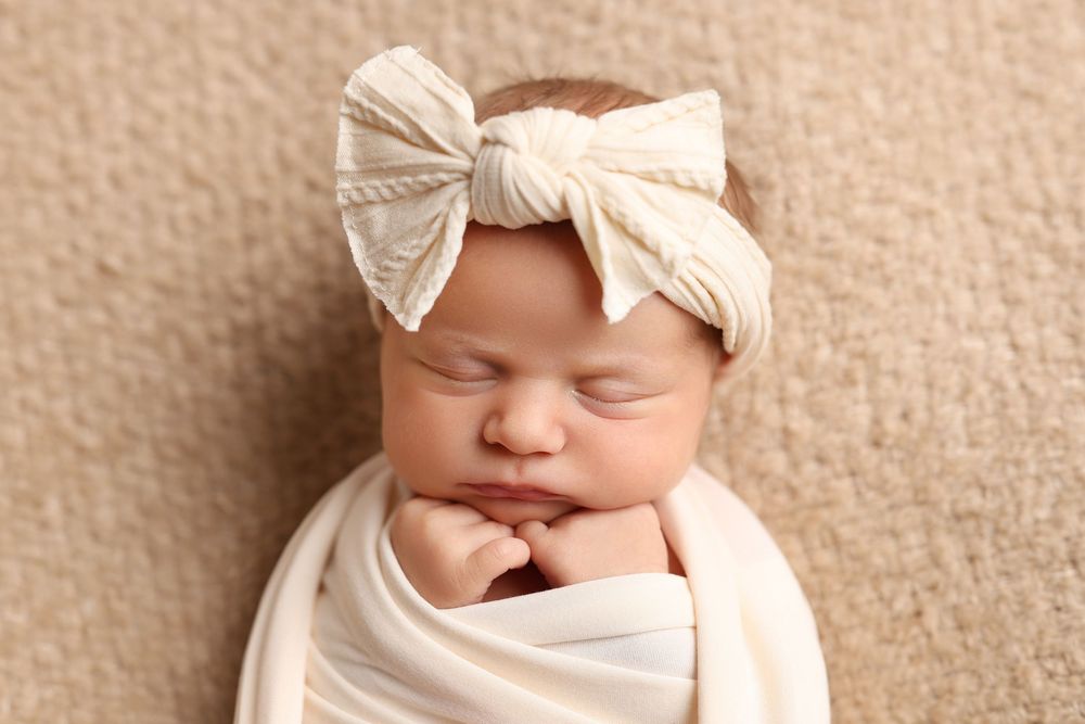 Sleeping newborn baby swaddled in cream wrap wearing a white bow headband on a beige background in Covington, GA