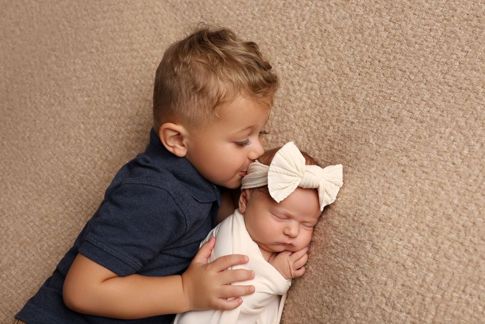 A young boy gently kisses his sleeping newborn sister wrapped in white, wearing a pink bow headband in Covington, GA