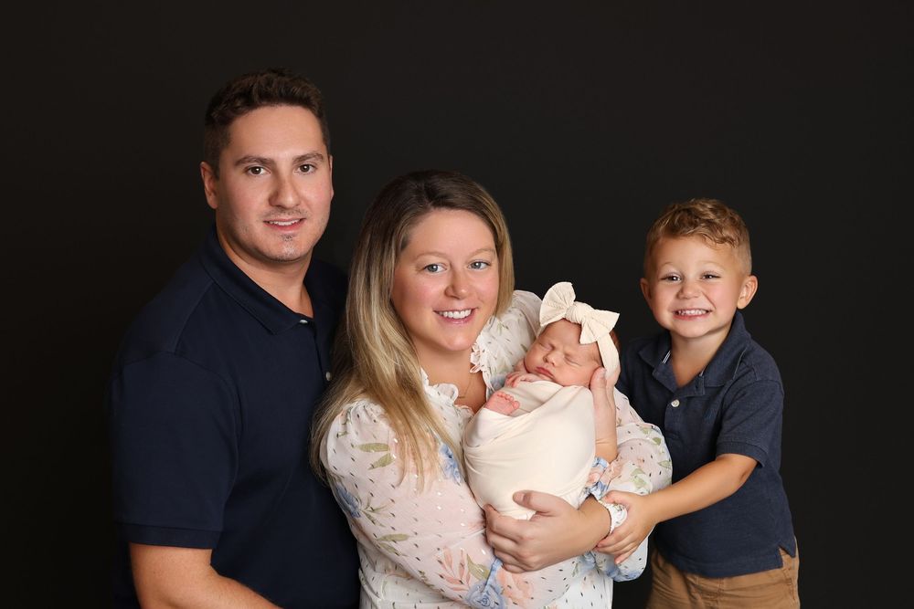 Family of four posing for a professional portrait, parents holding newborn baby and young boy, against dark background in Covington, GA
