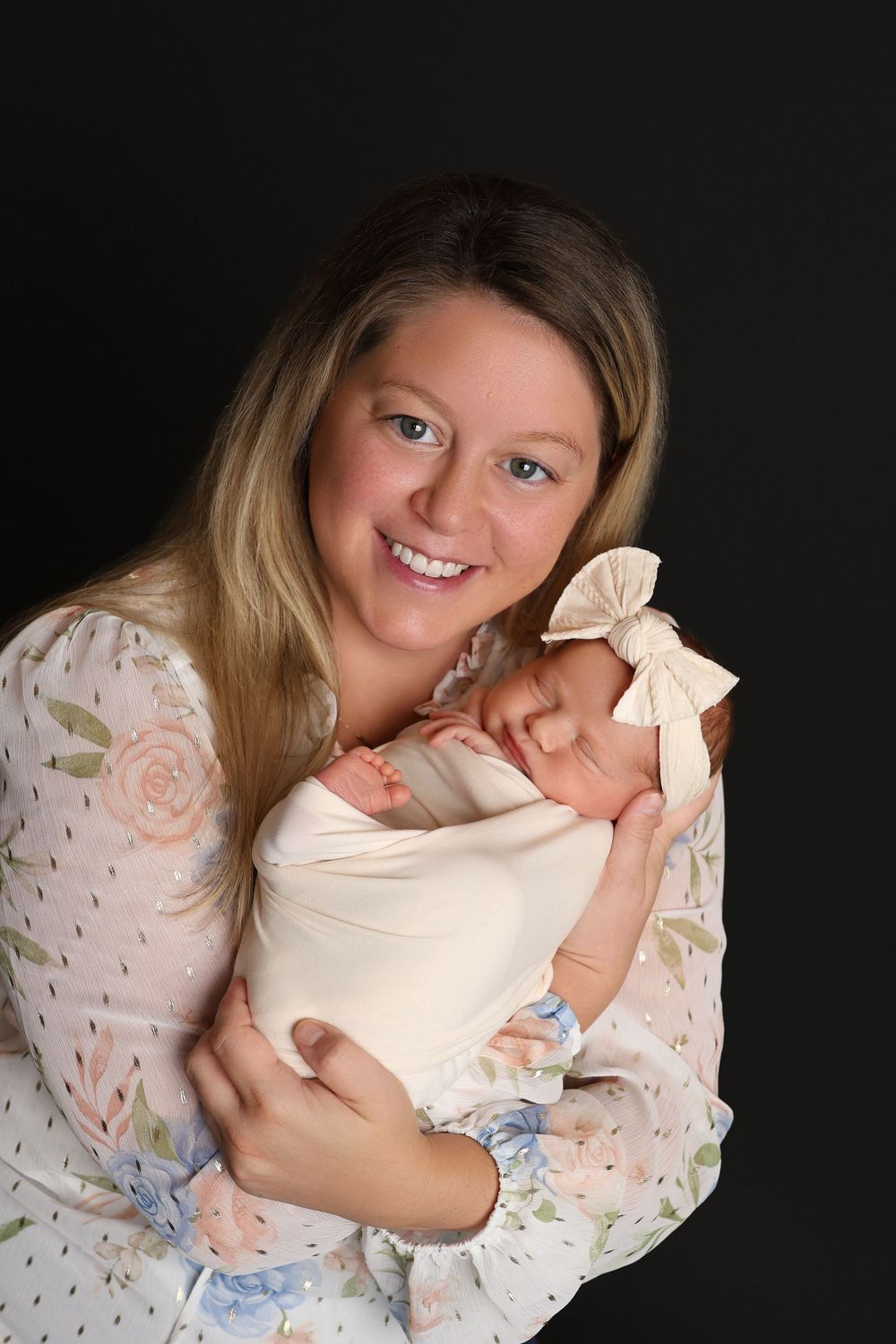 Smiling mother with blonde hair holds sleeping newborn baby girl wrapped in cream swaddle with bow headband in Covington, GA