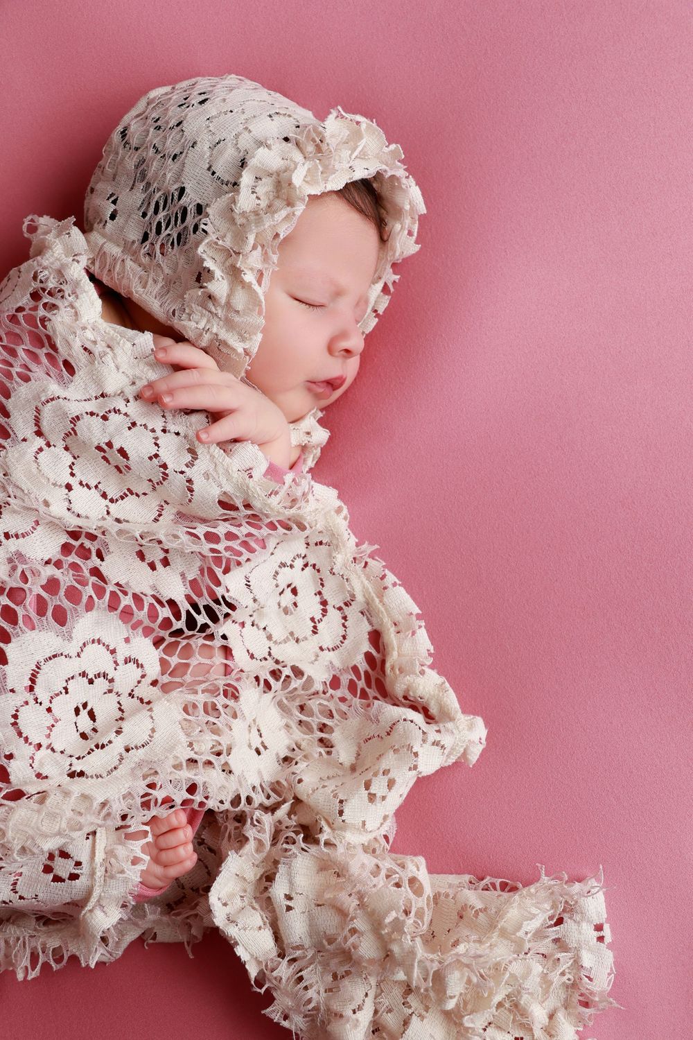 Sleeping newborn baby wrapped in white lace blanket and bonnet on pink background. photographer covington, ga