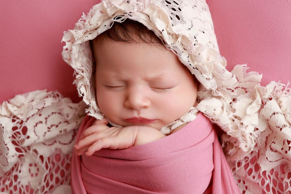 Sleeping newborn baby wrapped in pink swaddle wearing white lace bonnet on pink background. photographer covington, ga