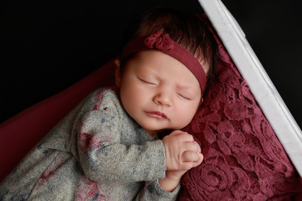Peacefully sleeping newborn in gray floral outfit with burgundy headband in white crib. photographer covington, ga