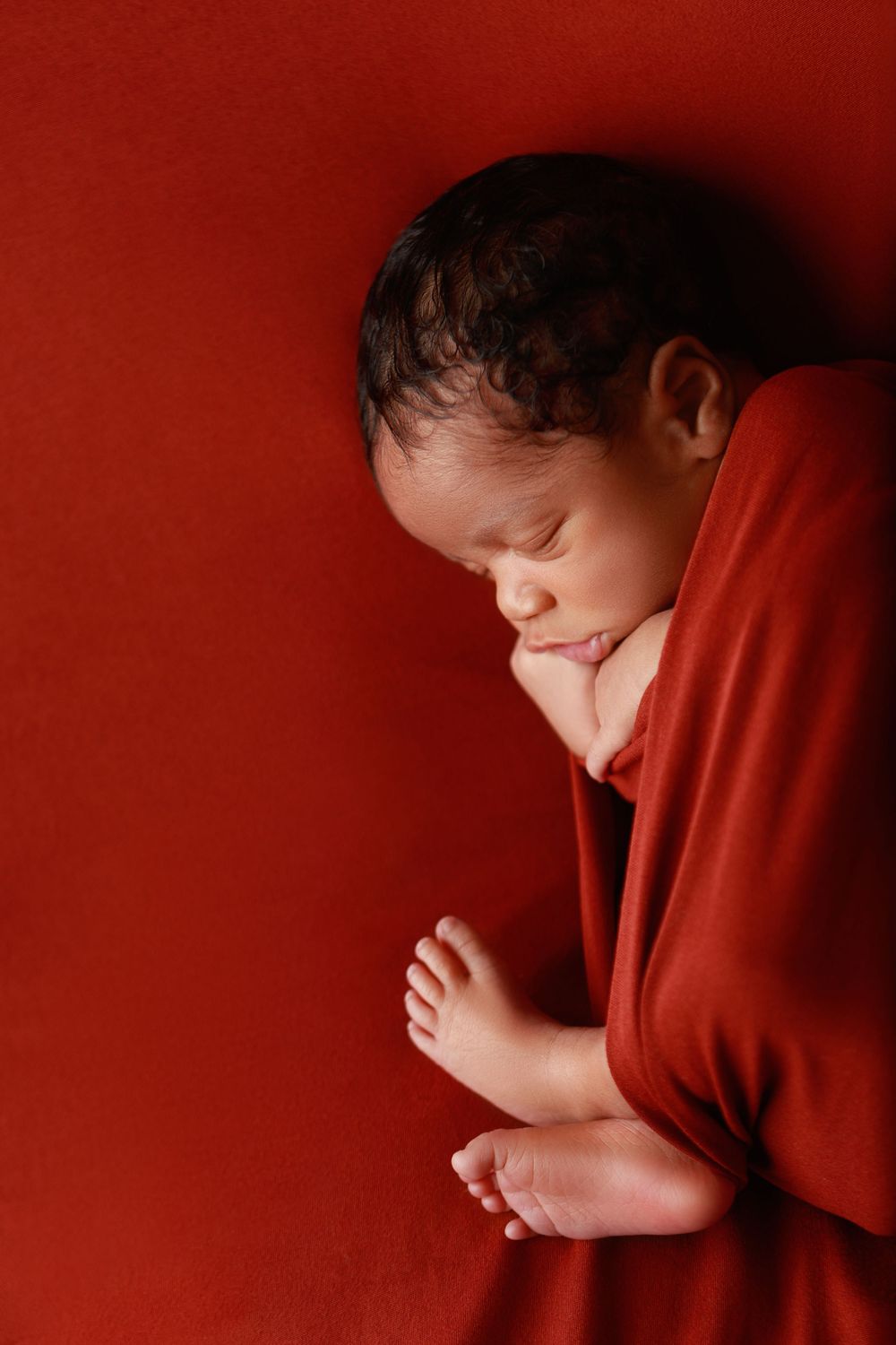Sleeping newborn baby wrapped in red swaddle with tiny feet visible on red background. photographer covington, ga