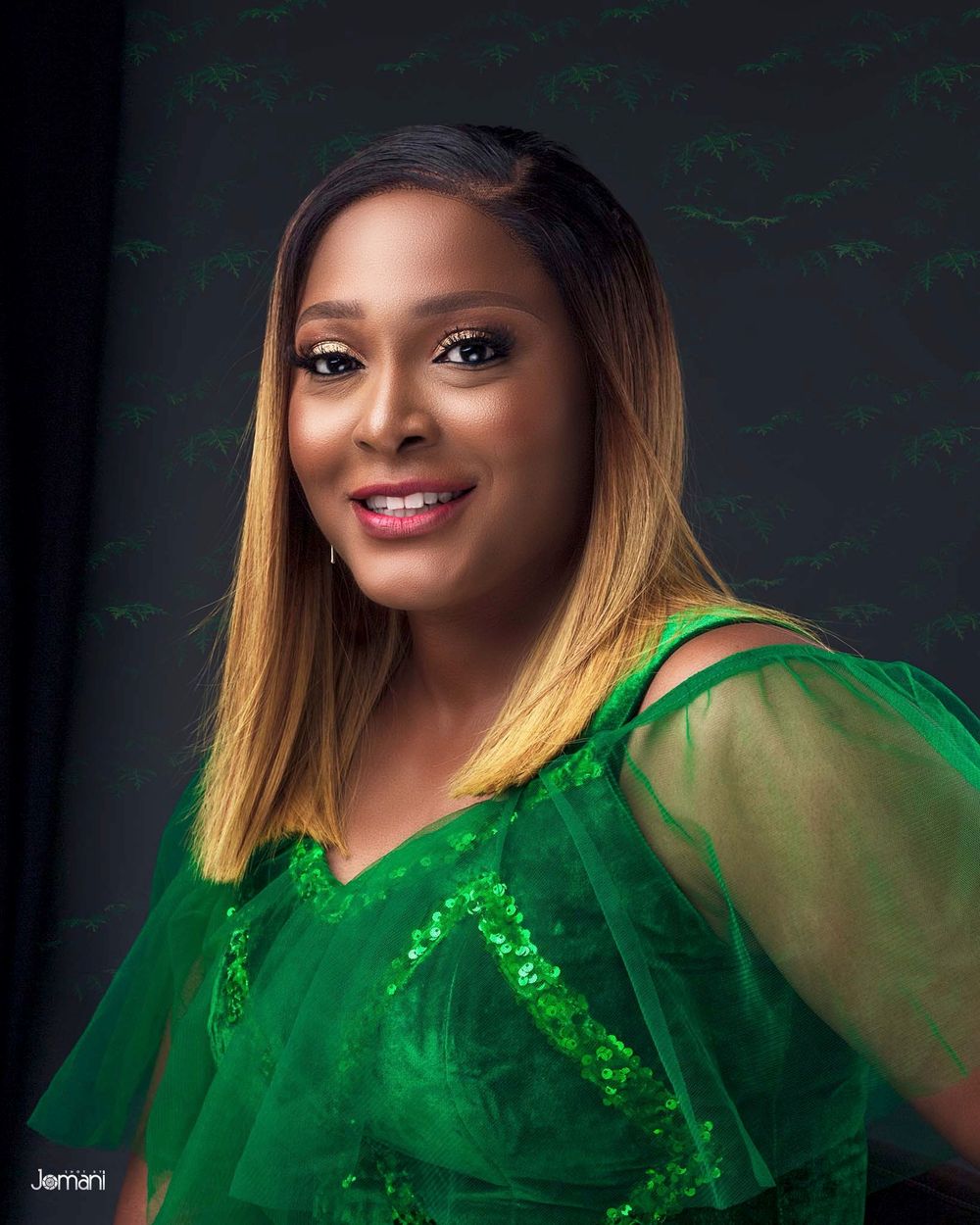 Smiling woman with blonde ombre hair wearing sparkling green sheer top in studio portrait.