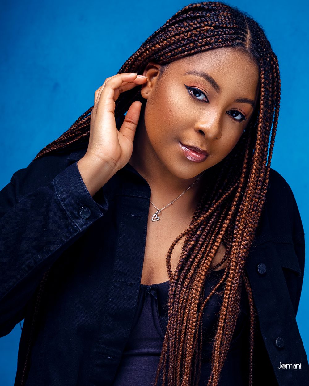 Woman with long brown box braids wearing a dark denim jacket poses against a blue background.