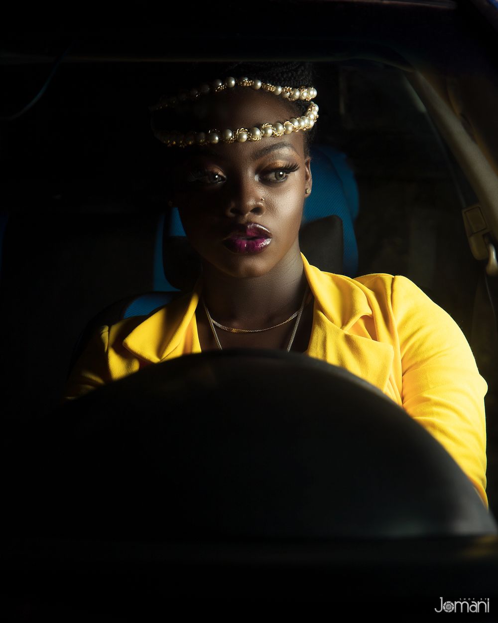 Woman with pearl headpiece in yellow jacket photographed through car windshield in dramatic lighting.