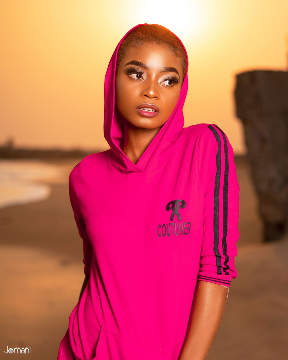 Young woman wearing bright pink hoodie with logo posing at sunset on beach.